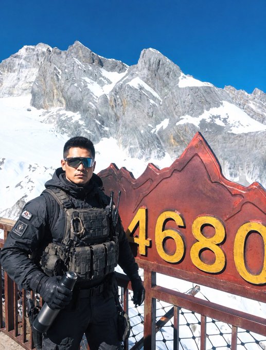 The image features a man standing next to a sign, possibly at the top of a mountain or near a bridge. The man appears to be dressed in military clothing, which suggests that he might be a soldier or a member of the military. He is holding a water bottle and wearing sunglasses, indicating that it could be a sunny day. The image also contains text elements such as "StandX" and "Top 100", possibly referring to an event or organization related to the man's military background.