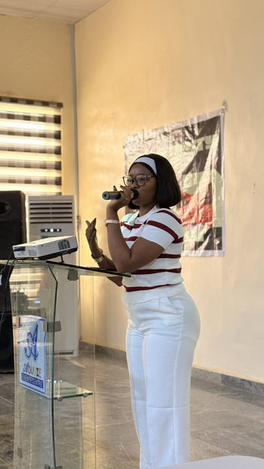 A woman with glasses, a headband, and a white and maroon striped shirt speaks into a microphone at a podium during the MarsChainDAO event in Abuja, with a banner reading "Mars Chain" visible behind her.