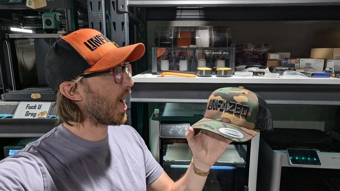 A man wearing an orange and black "UNFAZED" trucker hat excitedly holds up a camouflage "UNFAZED" snapback hat while promoting a giveaway, with a Bitcoin mining setup visible in the background alongside a humorous "Fuck U Greg" sign.