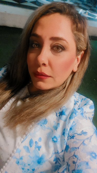 A selfie of a woman with long blonde hair wearing makeup and a white shirt with a blue floral pattern.