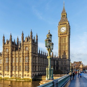 The iconic Big Ben clock tower and the Palace of Westminster in London, viewed from Westminster Bridge with pedestrians walking along the sidewalk.
