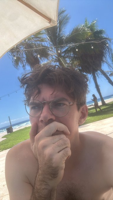 A shirtless young man with glasses takes a selfie outdoors under a beach umbrella, with palm trees and the ocean visible in the background.