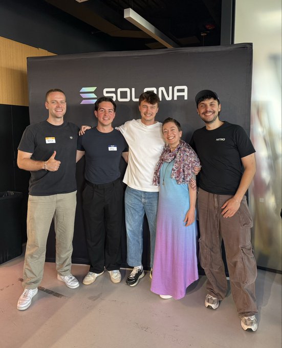 The image features a group of five people standing together in front of a black backdrop with the word "Solana" written on it. The individuals are posing for a photo, with one person holding their arm around another's shoulder as they all smile. They appear to be attending an event or gathering related to Solana, possibly a career day or a meeting.