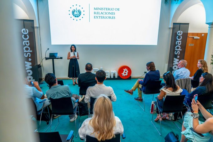 A woman speaks at a podium during an event at the "Ministerio de Relaciones Exteriores" with a Bitcoin logo and "ow.space" banners displayed, as El Salvador opens an Honorary Consulate in Lugano to strengthen Bitcoin ties with Switzerland. Audience members sit attentively, listening to the presentation in a formal diplomatic setting.