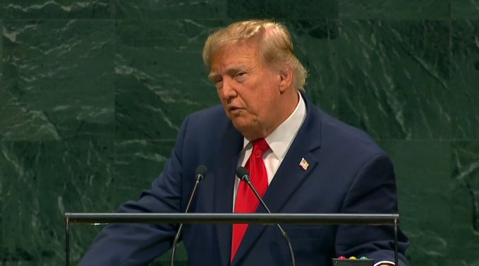 Donald Trump, wearing a navy suit with a red tie and American flag pin, stands at a podium with microphones against a green marble backdrop.