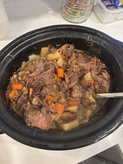 A slow-cooked beef stew with shredded meat, chunks of potatoes, carrots, and barley in a dark broth, served in a black ceramic bowl with a spoon resting inside.