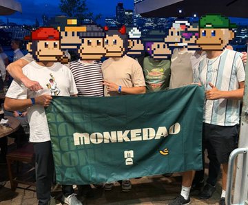 A group of five people posing indoors at night, their faces covered by cartoon monkey emojis, holding a large green flag with the text "MONKEDAO".