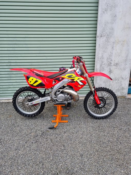 A red Honda dirt bike with the number 97 sits on an orange stand in front of a green roll-up door.