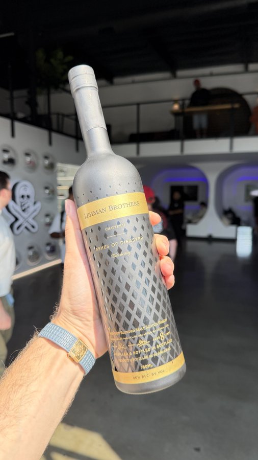 A hand holding up a silver metallic wine bottle labeled "LEHMAN BROTHERS CHAPTER 11 ASHES OF DISASTER 750ML 40% ALC. BY VOL." in an industrial-style interior setting.