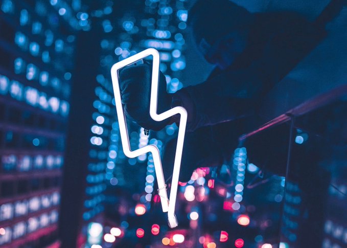 A person holds a glowing neon lightning bolt light against a background of blurred blue and pink city lights at night.
