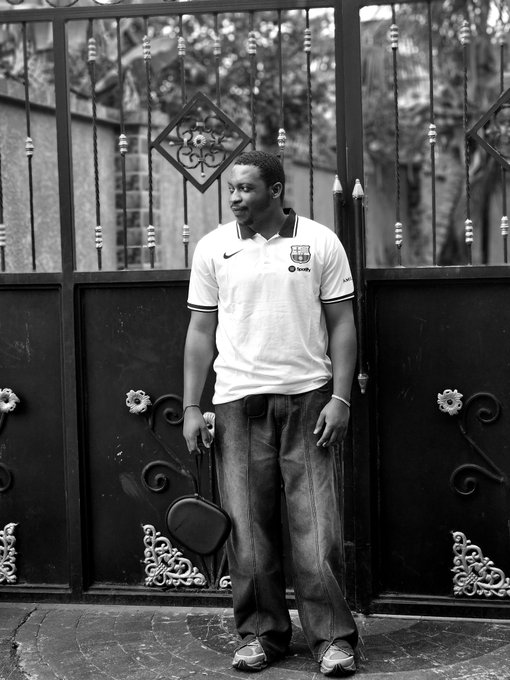 A black and white portrait shows a young man wearing a white Nike polo shirt and jeans, standing in front of an ornate metal gate while holding a small bag.