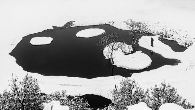 An aerial black-and-white view of a partially frozen lake in a snowy landscape, showing snow-covered islands, bare trees, and a small figure walking across the ice.