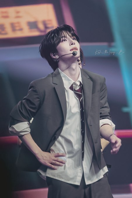 A young male performer, likely Xinlong (씬롱/鑫隆/贺鑫隆) of ALD1 (알디원/ALPHADRIVEONE), is captured mid-performance wearing a headband microphone, a white collared shirt with rolled sleeves, a loosened striped tie, and a grey blazer, with his head tilted upward on a stage lit with blue and red hues. The watermark "kst_theboy_01" is faintly visible in the upper right, and the post's excitement about a "bookshelf" pose suggests this striking stance with hands on hips is the moment being celebrated.