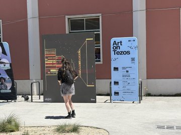 The image features a woman standing next to an art exhibit, looking at a large screen that displays artwork from Tezos. The woman has her back turned towards the camera, giving us a clear view of the artwork she's observing. In addition to the woman, there are two other people in the scene - one person is located near the left side and another person can be seen on the right side of the image. 

The post includes various images such as an advertisement for Sinusoidalsnail, a picture of a box with a cat inside, and a picture of a bird. These images are scattered throughout the post in different sections, adding to its overall visual appeal.