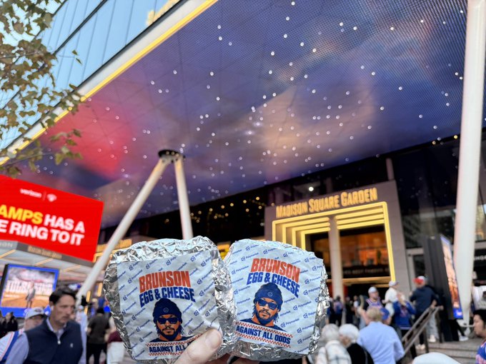 A fan holds two foil-wrapped “BRUNSON EGG & CHEESE” sandwiches outside Madison Square Garden, where many fans are gathered. The stadium-branded food connects to the post’s praise of Polymarket’s sports-fan engagement by highlighting a game-day twist on a celebrity-named meal.