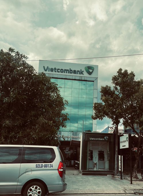 A silver van with license plate "92LD-00134" is parked outside a Vietcombank branch featuring a glass facade and a small ATM booth, likely taken by someone about to withdraw money from their Pay account. The image illustrates the Vietnamese poster's concern about the rush to withdraw funds and potential issues with held payments.