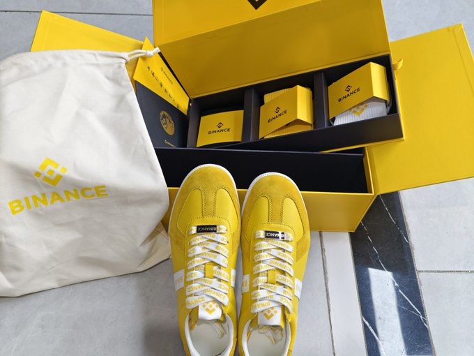 A pair of bright yellow Binance-branded sneakers is displayed next to their matching yellow shoebox and a Binance drawstring bag, with smaller Binance accessory boxes tucked inside the packaging. The image reflects the excitement in the caption about finally receiving the highly anticipated yellow Binance shoes, matching the enthusiasm for "元元" (Binance's mascot) merchandise.