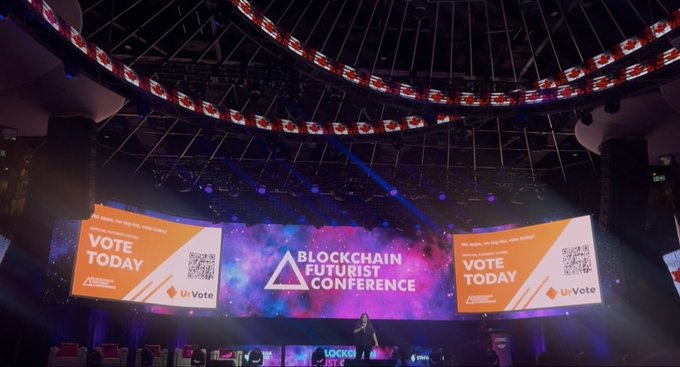A photo from inside a large conference venue displaying 'BLOCKCHAIN FUTURIST CONFERENCE' on stage screens with 'VOTE TODAY' messages, a speaker on stage, and elaborate lighting fixtures overhead.