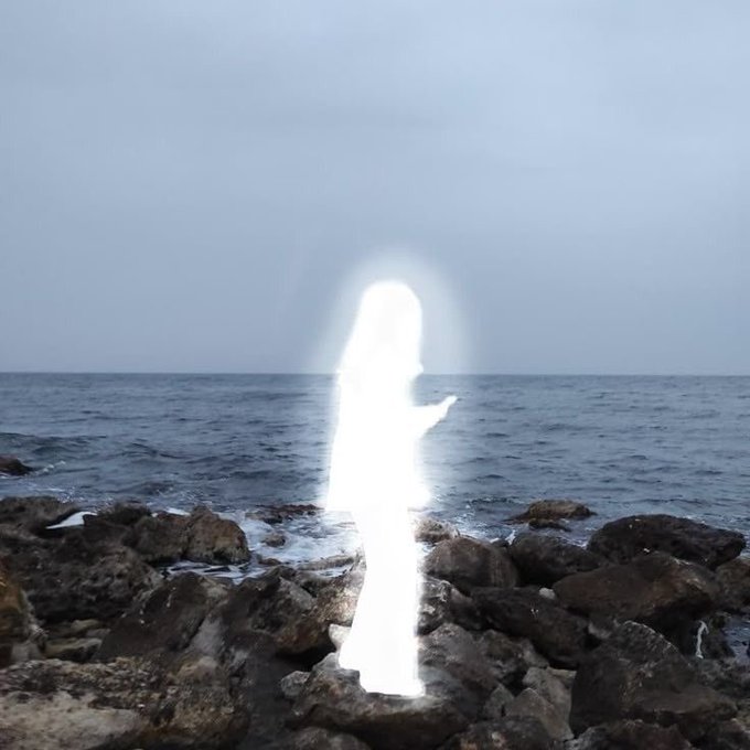 A glowing, ethereal white silhouette of a person stands on dark, jagged rocks by a calm ocean under a hazy, overcast sky.