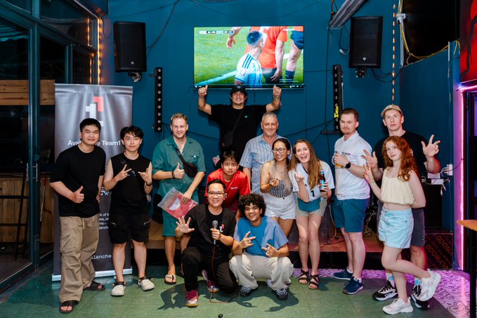 A diverse group of people posing together at what appears to be a sports bar or event venue, with a TV showing sports footage in the background and an 'HF' banner visible.