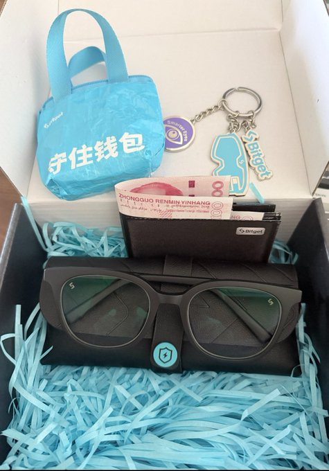 The image features a box containing various items, including sunglasses, a wallet, and some money. The box also contains a keychain with a picture of an eye on it. The post includes text that reads "贝姐和Bitget共同提醒你：收到的所有 Bitget 官方邮 专属防钓鱼码", which suggests that the items in the box are related to a product or service provided by Bitget. The post also mentions "杜绝对AI助手的过度授权钱包" and "不扫来历不明的二维码", which could be related to the wallet or keychain in the box.