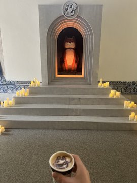 A scene showing a person holding a bowl while facing a cat sitting inside an altar-like structure surrounded by lit candles.