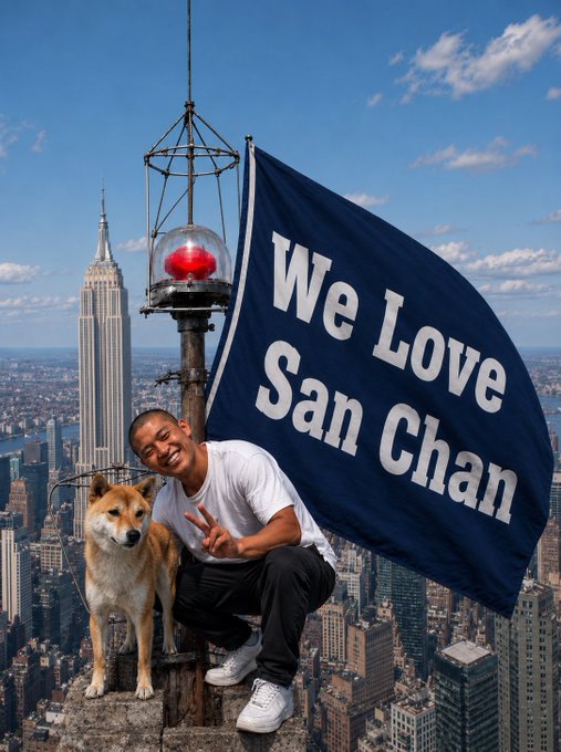 A young man crouches next to a Shiba Inu dog atop a skyscraper with a "We Love San Chan" flag, with the Empire State Building visible in the background cityscape.