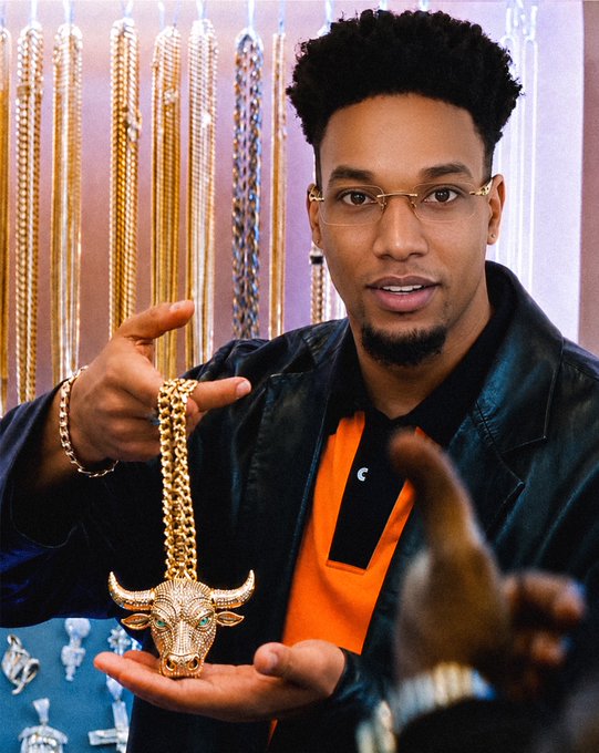 A man with glasses and a goatee, wearing a black jacket over an orange shirt, proudly displays a large gold bull pendant necklace against a backdrop of hanging gold chains. The image, captioned "this is how we win. $ANSEM," conveys a bullish sentiment toward the meme coin $ANSEM, with the bull symbol representing market optimism and upward momentum in crypto.