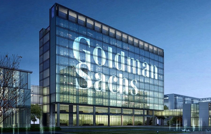 The Goldman Sachs headquarters building is shown at dusk, with the company's name illuminated in large letters across the glass facade of the modern office tower. The image directly relates to the post as it depicts the Goldman Sachs corporate building, whose bank has revised its forecast to expect a 25 basis point Fed rate hike at the upcoming September meeting.