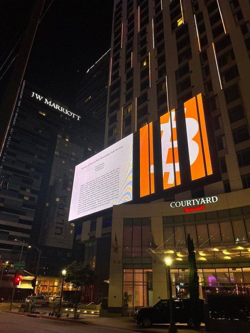 In the image, there are two buildings located next to each other - one is a tall building with a billboard featuring the Bitcoin symbol, while the other is a shorter building that has a sign for the Courtyard hotel. The Bitcoin White Paper is displayed on a large screen in front of this shorter building, which seems to be an advertisement or promotion related to Bitcoin.