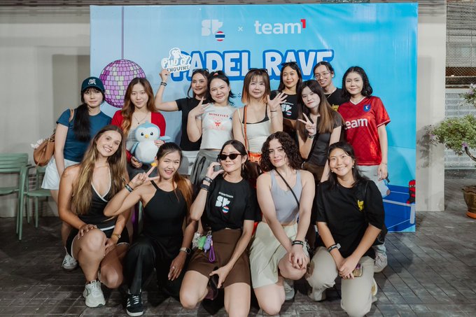 A group of women posing for a photo in front of a blue banner that reads 'PlayStation x team1 PADEL RAVE'. Some women are holding stuffed toys, and a person in an owl mascot costume is visible in the back.