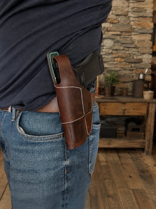 A person wearing jeans and a dark shirt carries a brown leather holster on their hip with a smartphone and what appears to be a Glock handgun tucked inside.
