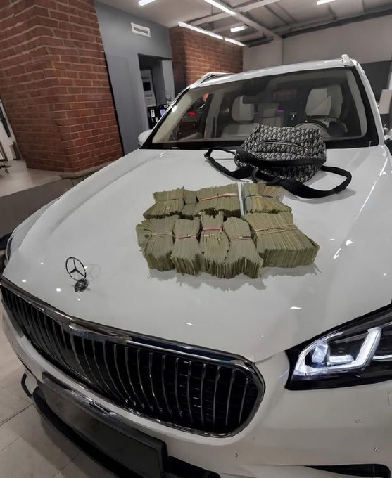 A white Mercedes SUV in a showroom has stacks of cash bound with rubber bands and a patterned duffel bag displayed on its hood.