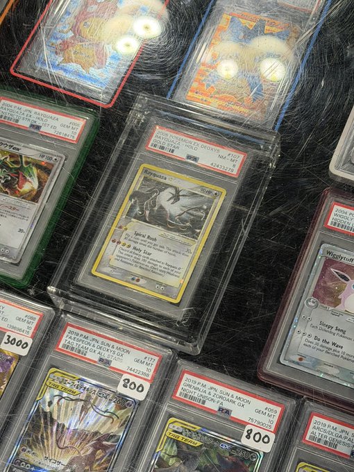 A "oh my god" moment unfolds over a graded PSA 7 Rayquaza Neo Holo (2002 Japanese) Pokemon card, marked down to just $300—a shockingly low price for such a rare holographic. Surrounding it are other notable PSA-graded cards, including a 2019 Japanese Sun & Moon Gengar & Weezing and a Teddiursa PSA 10, all part of a stunning collection.