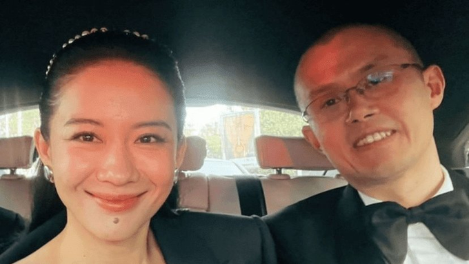 A smiling couple in formal attire (the woman in a dark dress with a headband and the man in a tuxedo with bow tie and glasses) pose for a selfie inside a car. The image illustrates the close partnership between Binance co-founders CZ and Yi He, who reportedly used the codenames "Homer" and "Marge" in internal meetings, fittingly matching their visibly affectionate, married-like dynamic in the photo.