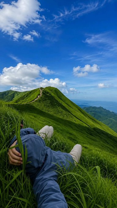 A scenic photograph of a person in blue pants and white sneakers lying in tall green grass on a mountain ridge, gazing out at rolling green hills under a bright blue sky with scattered clouds.