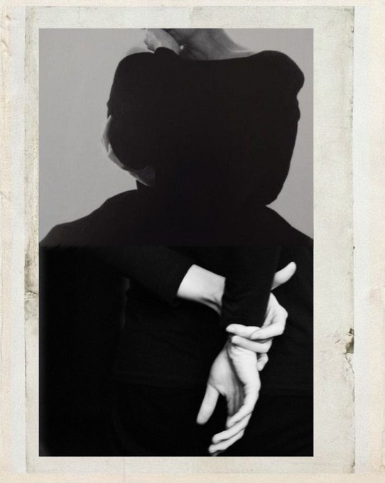 A black-and-white artistic photograph of a person in a dark off-the-shoulder top with their hands crossed behind their back, framed in a textured cream border.