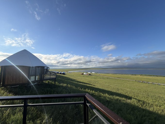 A scenic landscape photograph taken from a wooden balcony, showing a white geodesic dome lodge on the left, overlooking green grasslands that stretch toward a body of water under a blue sky with scattered clouds.