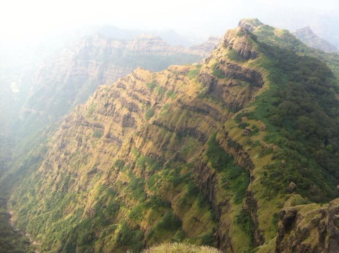 A scenic landscape view of a lush green mountain ridge with layered rocky cliffs, shrouded in mist and hazy sunlight.