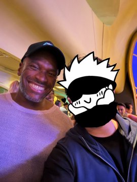 A selfie of a man in a dark cap and gray sweater smiling next to another person whose face is covered by a Kakashi (Naruto) mask sticker.