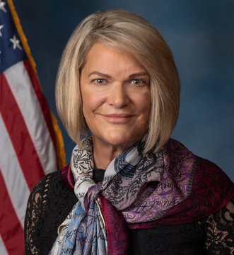 An official portrait shows Senator Cynthia Lummis standing in front of an American flag, wearing a patterned scarf and dark blazer with a slight smile. The image accompanies her quote advocating for the Clarity Act, a pro-Bitcoin and digital asset regulatory framework she promotes as a "game-changer" for building and growing crypto businesses in the U.S.