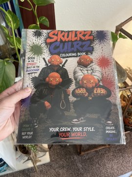 A hand holds up a copy of 'Skullz Guildz Colouring Book' featuring cartoon skulls in streetwear, with text reading 'YOUR CREW. YOUR STYLE. YOUR WORLD.'