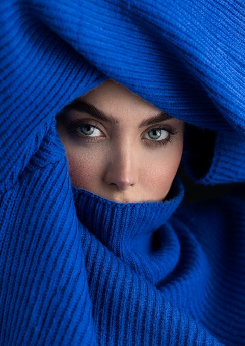 A striking portrait of a woman with piercing light eyes, wearing a vibrant blue ribbed knit garment that covers her head and wraps around her neck.