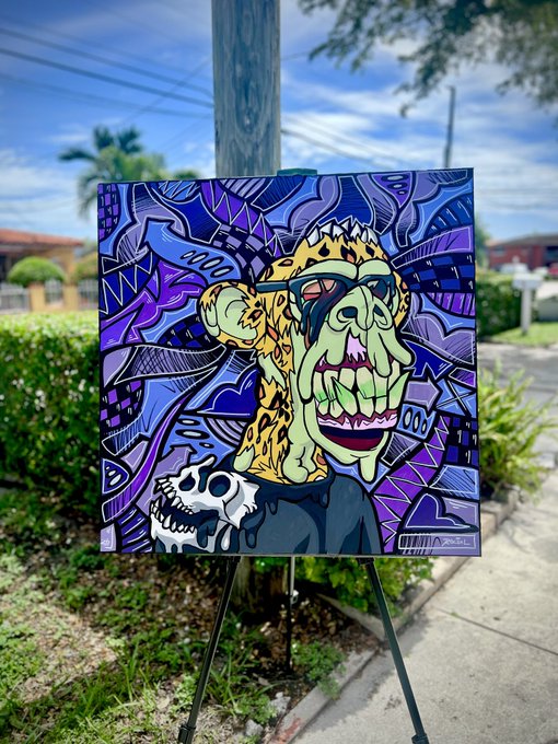 The image features a large painting of a monkey with a face painted on it, sitting on top of a wooden easel outdoors. The monkey has a unique design on its face that resembles a mask or a crown, giving it an artistic and whimsical appearance. The painting is displayed in front of a house, adding to the overall ambiance of the scene. The post accompanying this image reads: "Gm, y'all! I wanted to add some positivity to the timeline today—I just finished this 30x30 mutant ape canvas art piece for the one and only @hazy_purps. Love the different shades of purple—this one came out amazing. If anyone would love an original Roc Sol canvas, DM me for".