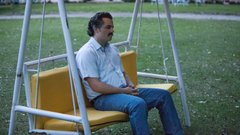 A still from a video or film showing a man with a mustache sitting on a yellow swing in a grassy area, looking off to the side.