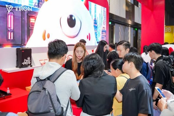 A crowd of people gathers at a TRON branded promotional booth featuring a large cartoon mascot, with the TRON logo visible in the upper left.