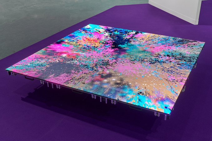 A vibrant, multicolored abstract painting on a square platform displayed on a purple floor, featuring splashes and splatters of pink, blue, yellow, and black, evocative of the creative and emotional NFT minting process described in the accompanying post.
