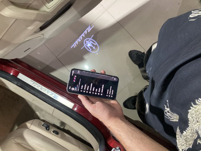 A person holds a smartphone displaying the audio equalizer app "Equalizer," while a Maserati logo is illuminated on the floor nearby, fitting the post's call for a morning "Gm" to begin the grind in style.