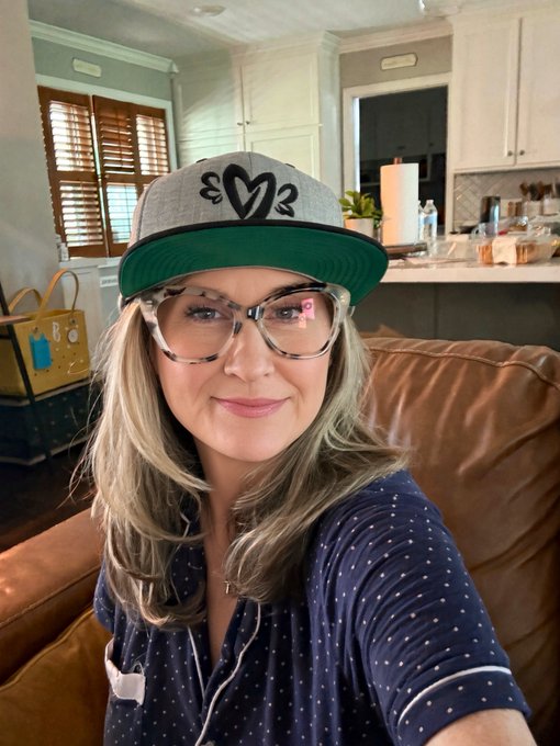 A woman with blonde hair and glasses wearing a navy polka-dot shirt and a gray and green 'heart' baseball cap takes a photo in a kitchen setting.