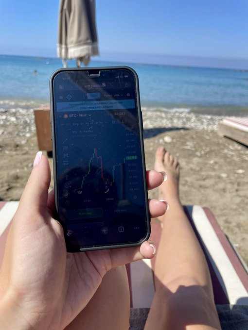 A person's hand holds a smartphone showing a BTC-PERP trading chart while sitting on a beach chair by the sea, with their bare feet visible in the background.